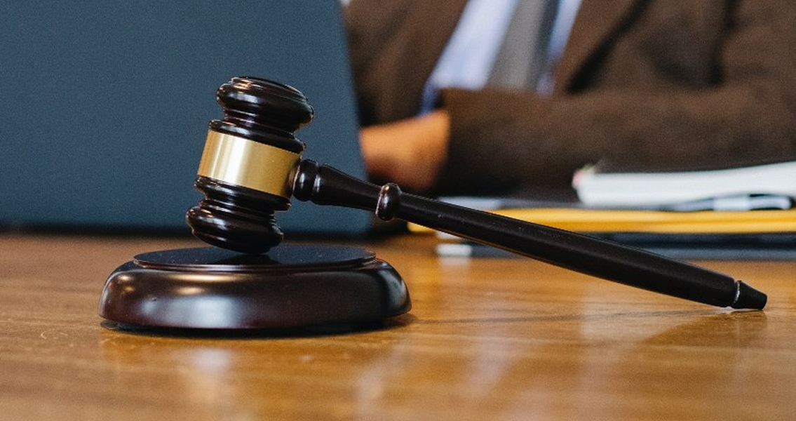 A wooden gavel rests on a judge's desk, with a blurred person in a suit using a laptop in the background. The scene conveys a legal and authoritative tone.