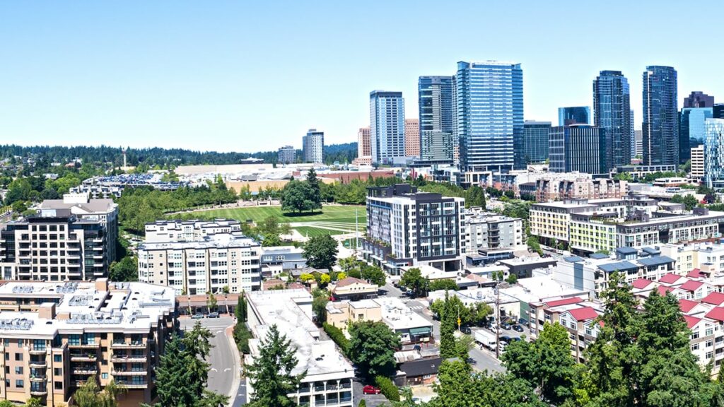 Aerial view of a cityscape with modern skyscrapers in the background, residential buildings in the foreground, and lush green areas under a clear blue sky.