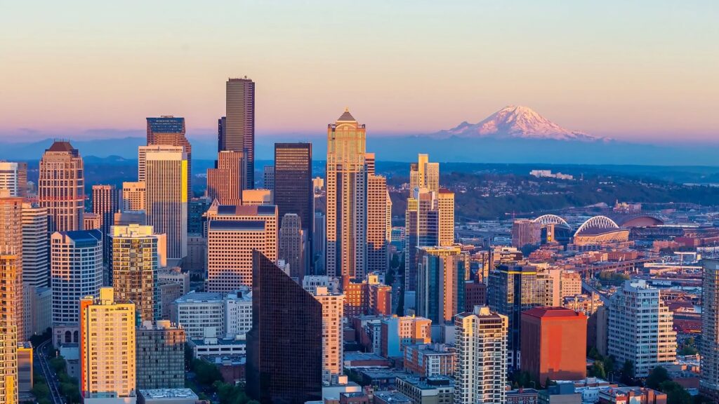 Seattle cityscape at sunset with skyscrapers bathed in warm light. Mount Rainier looms in the background under a soft pink sky, evoking tranquility.