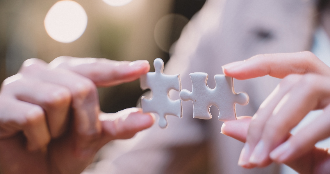 Two hands, each holding a puzzle piece, move together to connect them. The image conveys teamwork and problem-solving with a soft, focused background.