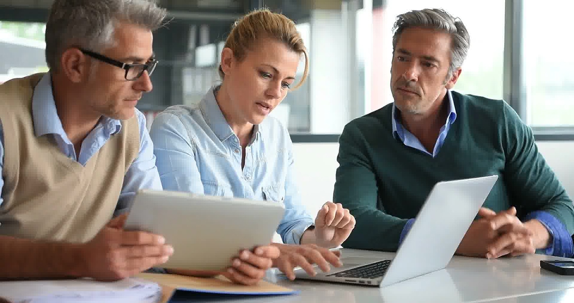 Three professionals collaborate at a table with a laptop and tablet. The atmosphere is serious and focused, reflecting a business meeting.