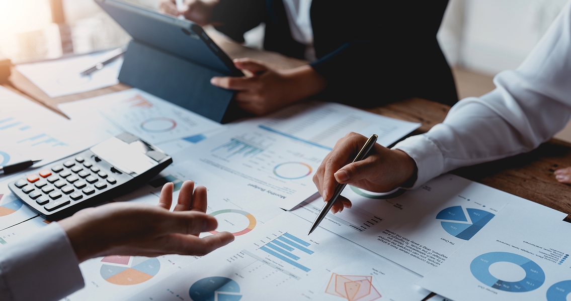 Business meeting scene with hands discussing charts and graphs. Papers cover the table, alongside a calculator and a tablet. Collaborative, focused atmosphere.