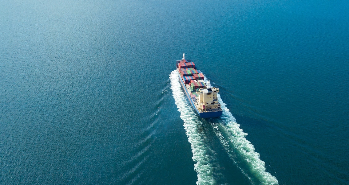 Aerial view of a large cargo ship moving through calm blue waters, its wake trailing behind. The ship is loaded with colorful shipping containers.