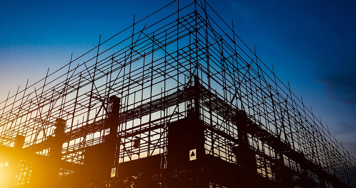 Silhouetted scaffolding frames a building under construction against a vibrant sunset and deep blue sky, conveying progress and anticipation.