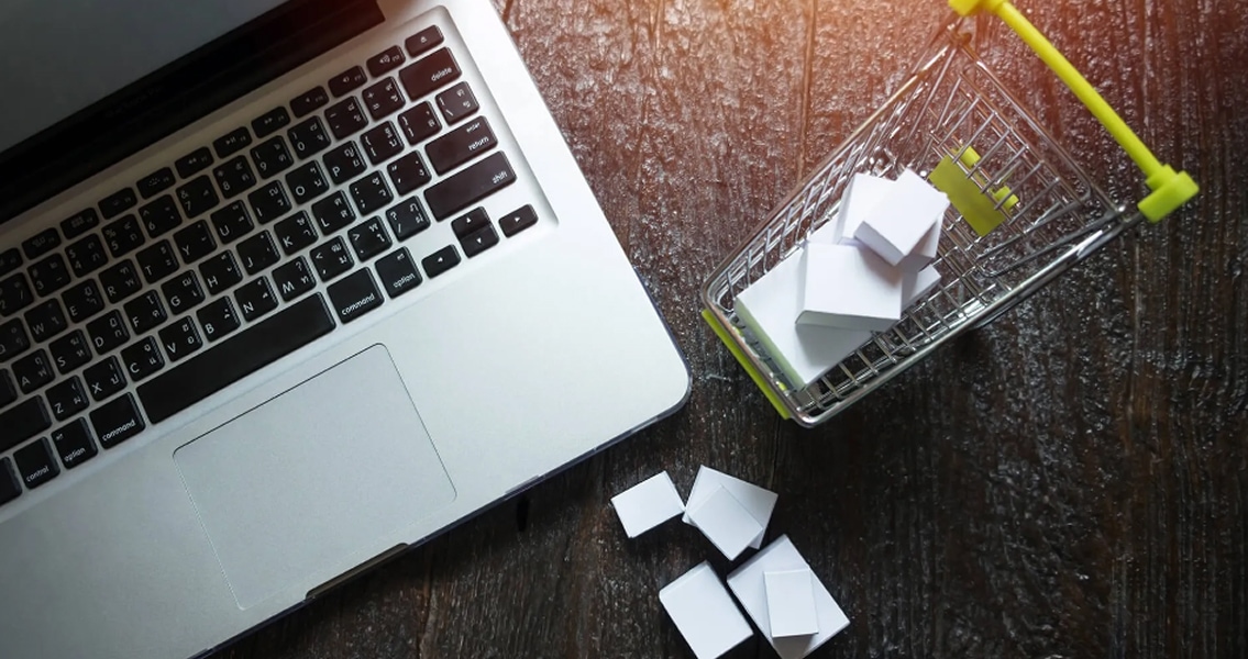 A laptop on a wooden table next to a miniature shopping cart filled with white boxes, symbolizing online shopping. Bright lighting creates a focused tone.