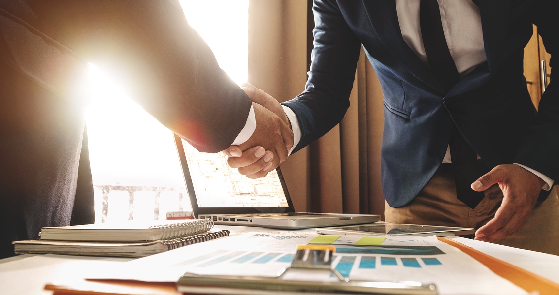 Two businesspeople in suits shake hands over a desk with documents and a laptop. Sunlight streams in, conveying a tone of agreement and success.
