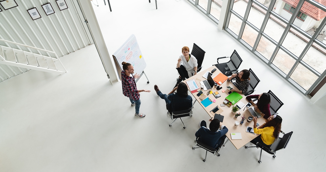 Overhead view of a diverse office team gathered at a table, with laptops and papers, as one team member stands and presents at a whiteboard. Bright and collaborative atmosphere.