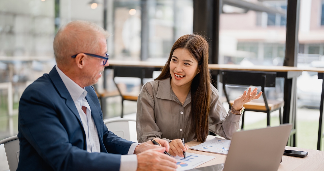 A senior man in a suit and glasses talks with a young woman in a professional setting. She's smiling, gesturing, papers with charts are on the table.