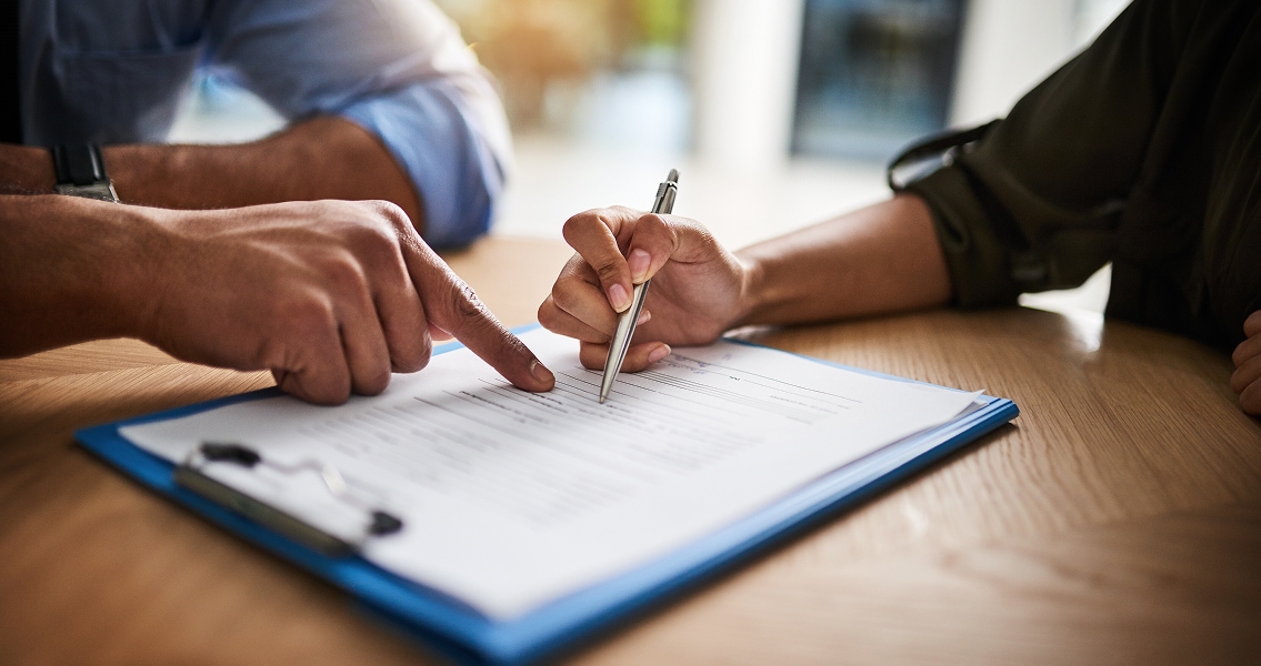 Two hands focus on a document on a table; one hand points while the other holds a pen, suggesting a discussion or review in a professional setting.