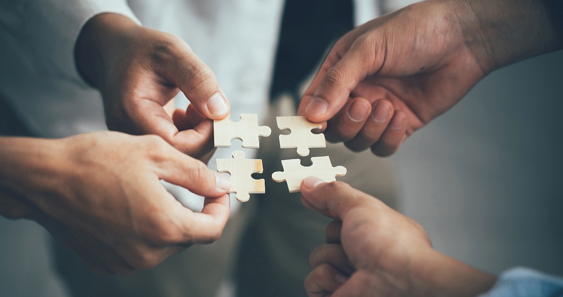 Four hands collaboratively hold interlocking puzzle pieces, symbolizing teamwork and unity. The scene conveys cooperation and shared problem-solving.