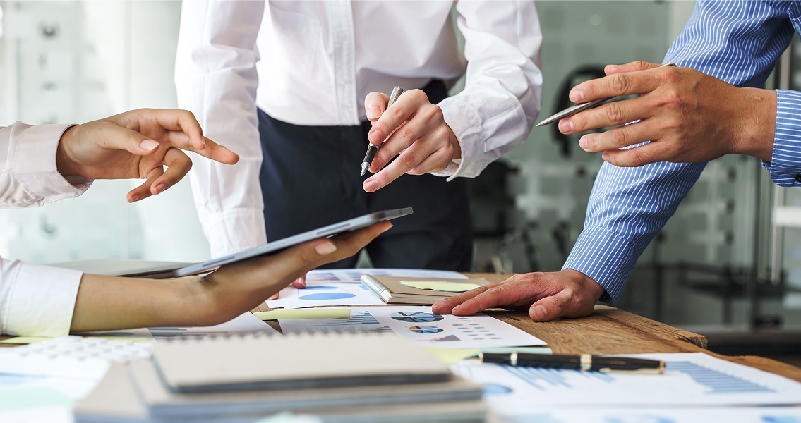 Four people collaborate over a wooden table covered with charts and documents. Hands gesture expressively as a tablet is used for discussion.