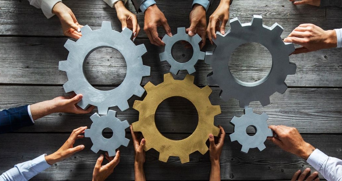 Multiple hands hold interlocking gears of varying sizes on a wooden surface. The scene conveys teamwork, collaboration, and problem-solving.