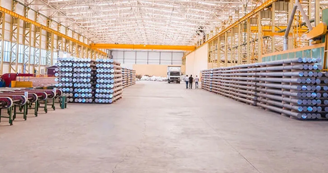 Vast warehouse interior with stacked metal tubes on both sides, illuminated by overhead lights. Two people and a truck appear small in the background.