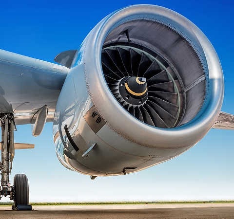 Close-up of a jet engine on an airplane wing, showcasing intricate turbine blades. The aircraft is on the tarmac under a bright blue sky.