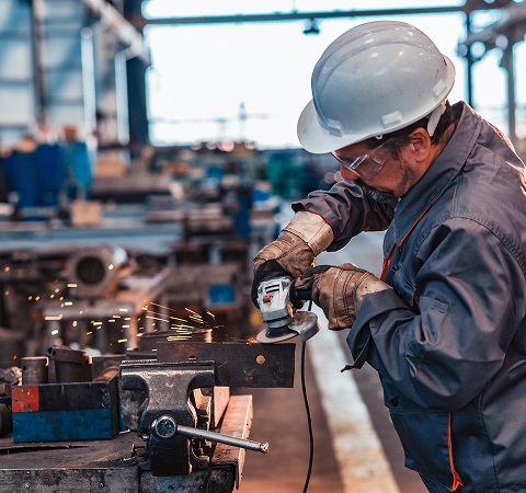 A worker in safety gear uses a grinder on metal, creating bright sparks, in an industrial factory setting, conveying focus and precision.