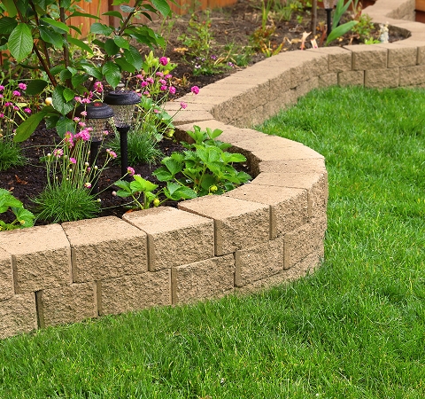 Curved tan stone garden border with vibrant green grass and pink flowers. Two solar lights add charm. The scene is peaceful and well-maintained.