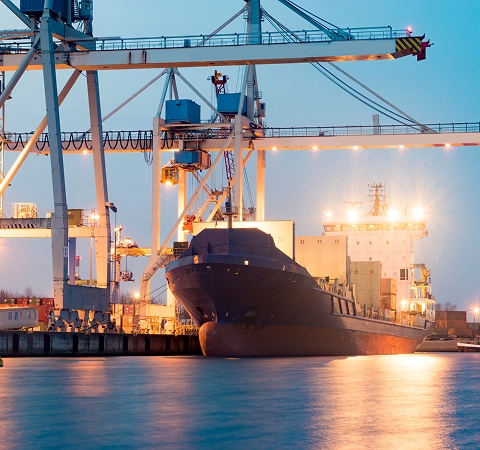 Container ship docked at a port in evening light, surrounded by illuminated cranes and reflecting on calm water, conveys industrial and tranquil mood.