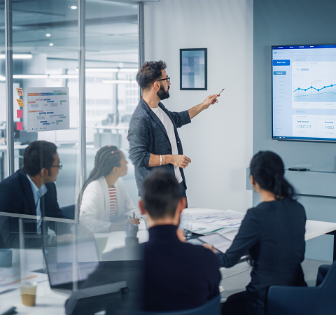 A man presents data on a screen to a diverse team in a modern office. Charts indicate business growth. The setting is professional and focused.
