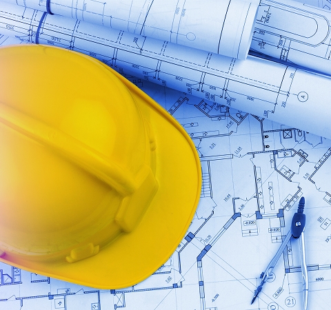 Yellow hard hat resting on detailed architectural blueprints, accompanied by a drafting compass, conveying themes of construction and design.