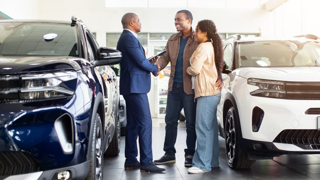 A smiling couple shakes hands with a car salesman in a showroom, standing between a blue and a white SUV. The atmosphere is friendly and positive.