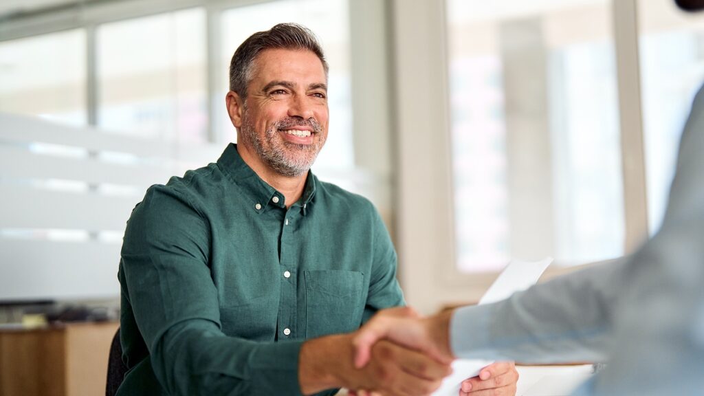 A man in a green shirt smiles warmly while shaking hands across a desk in a bright office setting, conveying a sense of agreement and positivity.