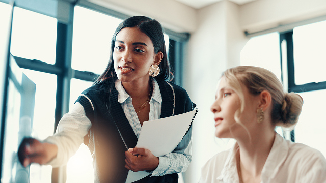 A woman with dark hair, holding papers, points at a screen, explaining something to a blonde woman. They seem focused, in a bright office setting.