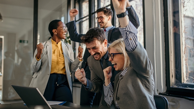 A diverse group of professionals in suits celebrate with raised fists and smiles in an office, suggesting a successful achievement or positive outcome.