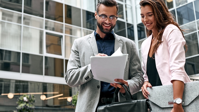 A man and woman smile while reviewing documents outside a modern glass building. Both appear professional and carry briefcases, conveying a business tone.