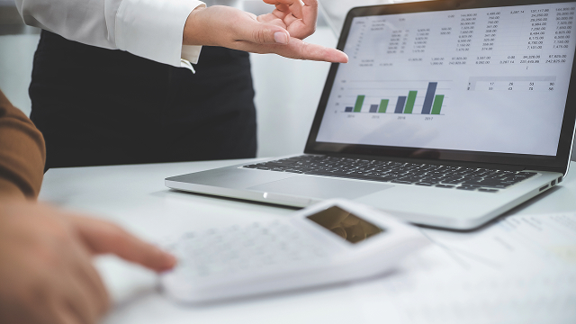 A person points at a laptop screen displaying data charts and graphs. Another individual uses a calculator. The scene conveys analysis and teamwork.