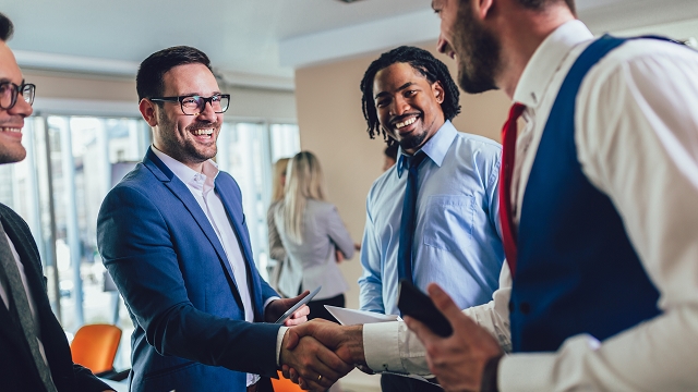 Group of professionals in business attire smiling and shaking hands in a bright, modern office. The mood is friendly and collaborative.