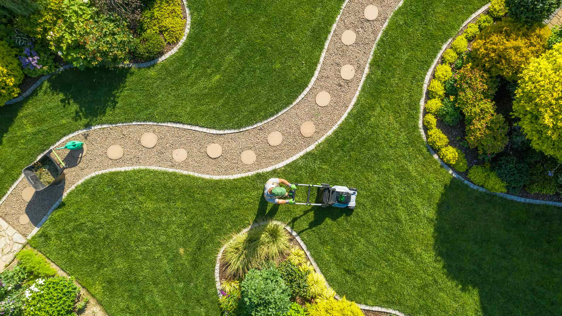 Aerial view of a landscaped garden with a curved gravel path and circular stepping stones. A person mows the lush green grass. Vibrant bushes surround the scene.