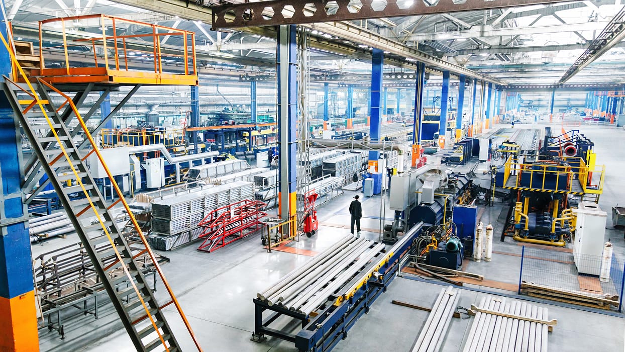 Industrial factory interior with high ceilings and bright lighting. Workers and machinery are visible, with rows of metal beams and equipment. The space feels busy and organized.