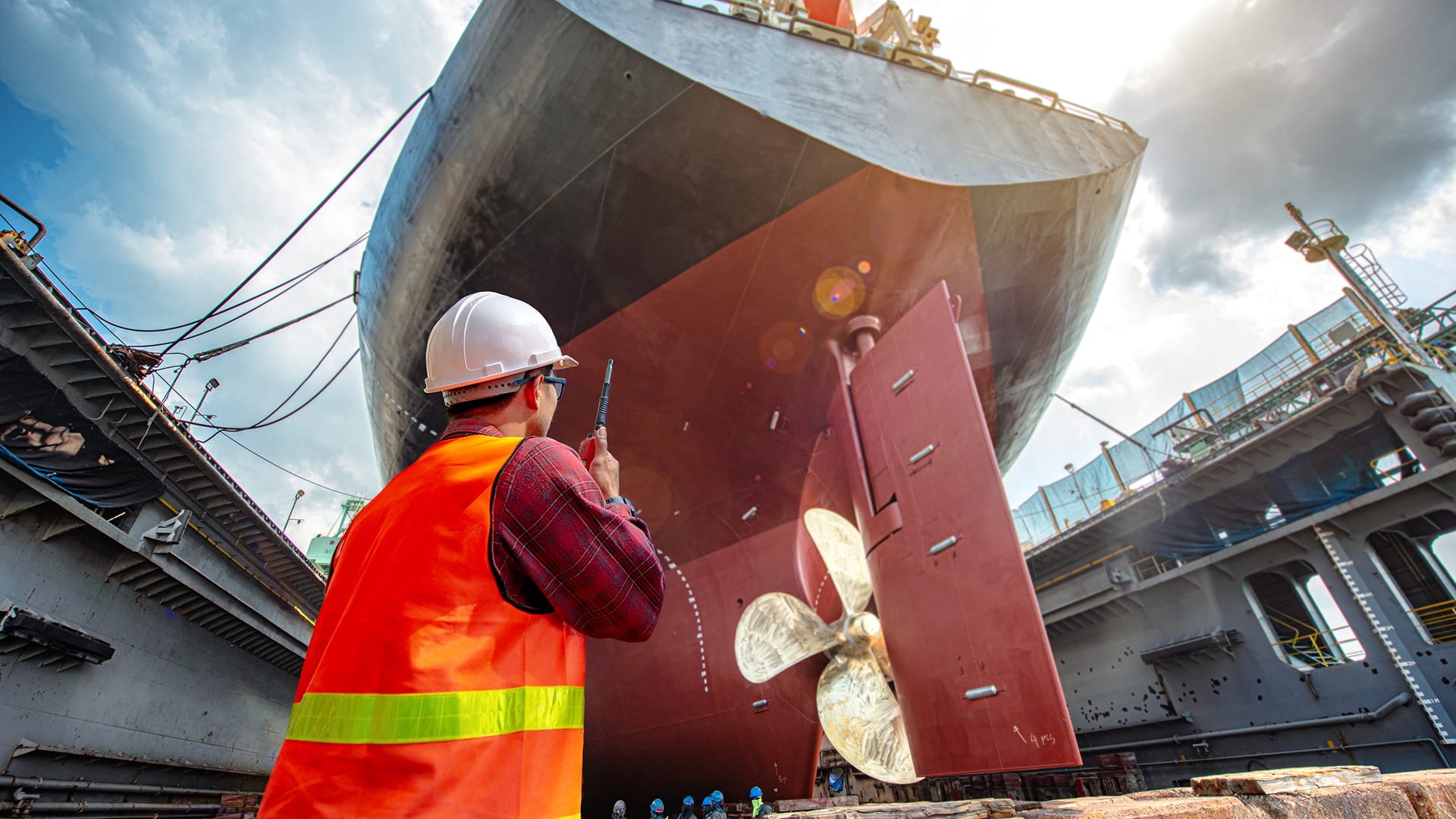 A worker in a reflective vest and hard hat inspects a massive ship's propeller in a dry dock. The scene conveys maintenance and focus under a bright sky.