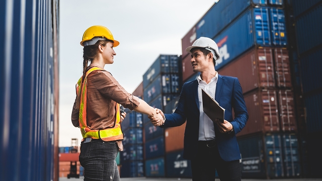 A woman in construction gear and a man in a suit with a hard hat shake hands, smiling, in front of stacked shipping containers in a busy port.
