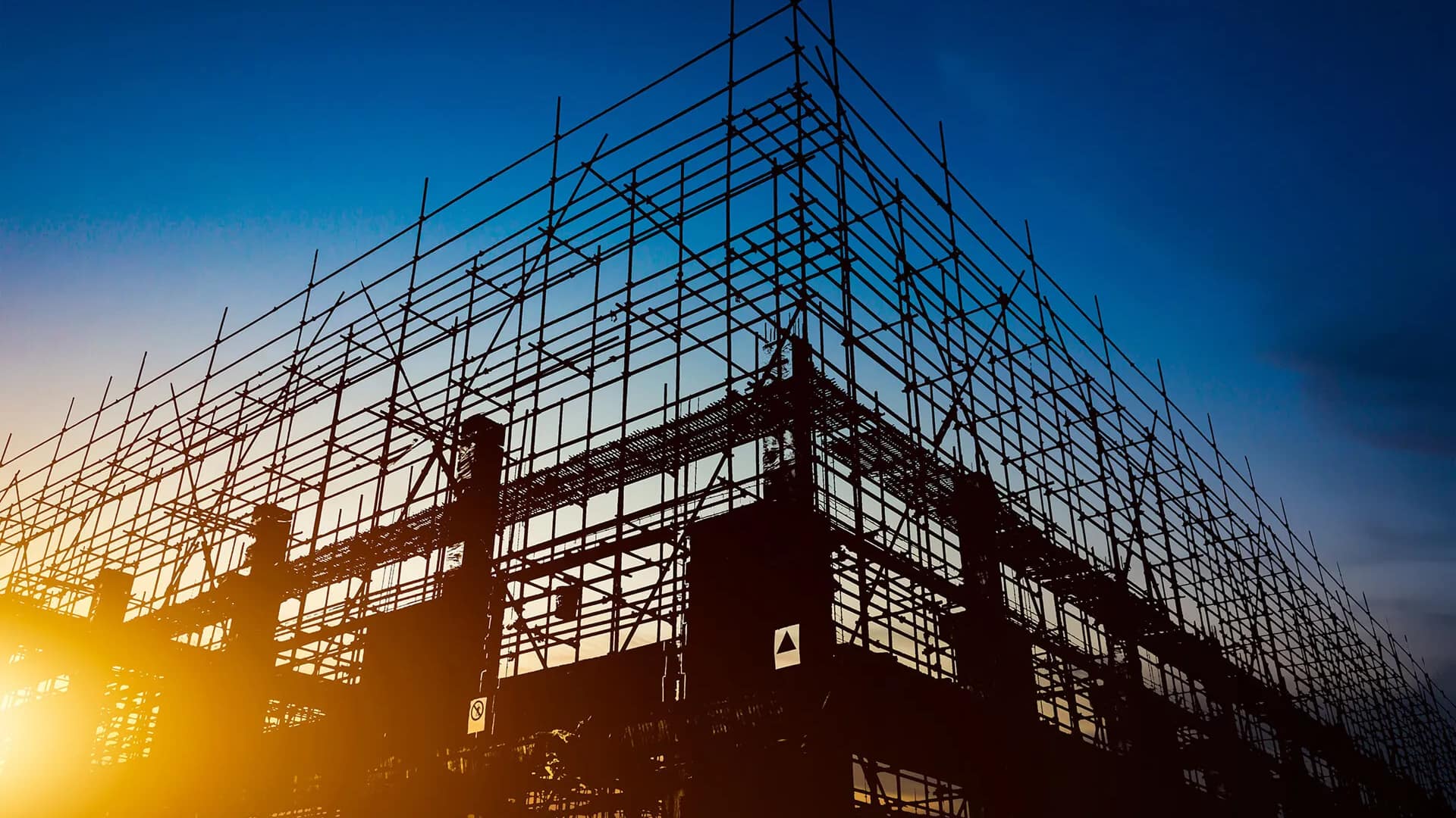 Silhouetted construction scaffolding against a twilight sky, with the warm glow of the setting sun. The image conveys a sense of progress and resilience.