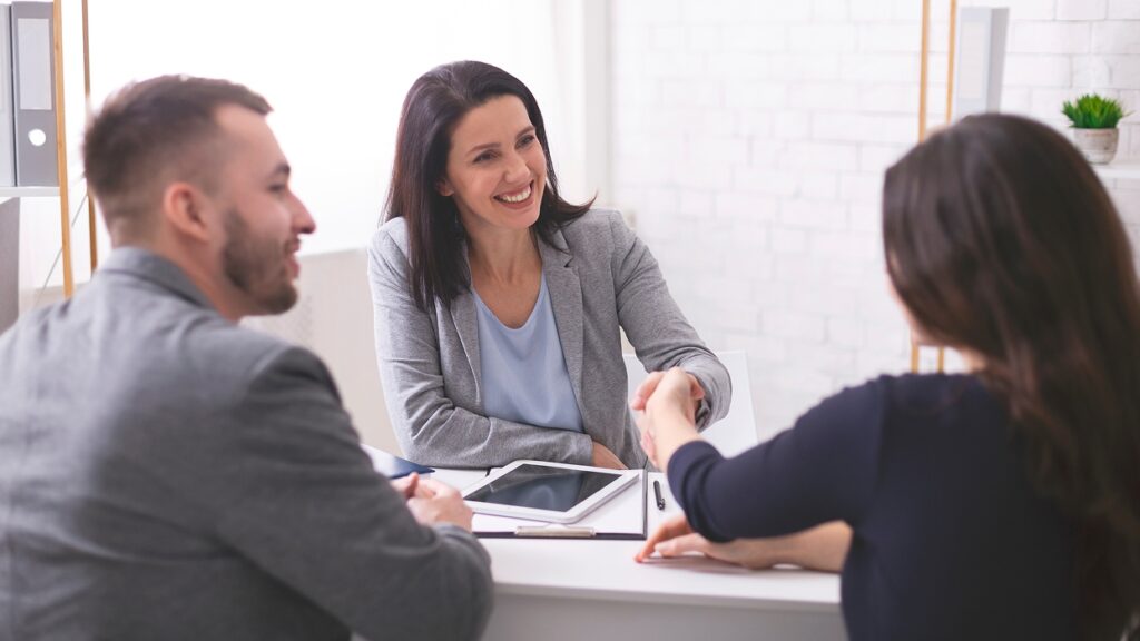 Three people in a professional setting smile as a woman in a gray blazer shakes hands with another woman. A man sits beside them, conveying a positive tone.