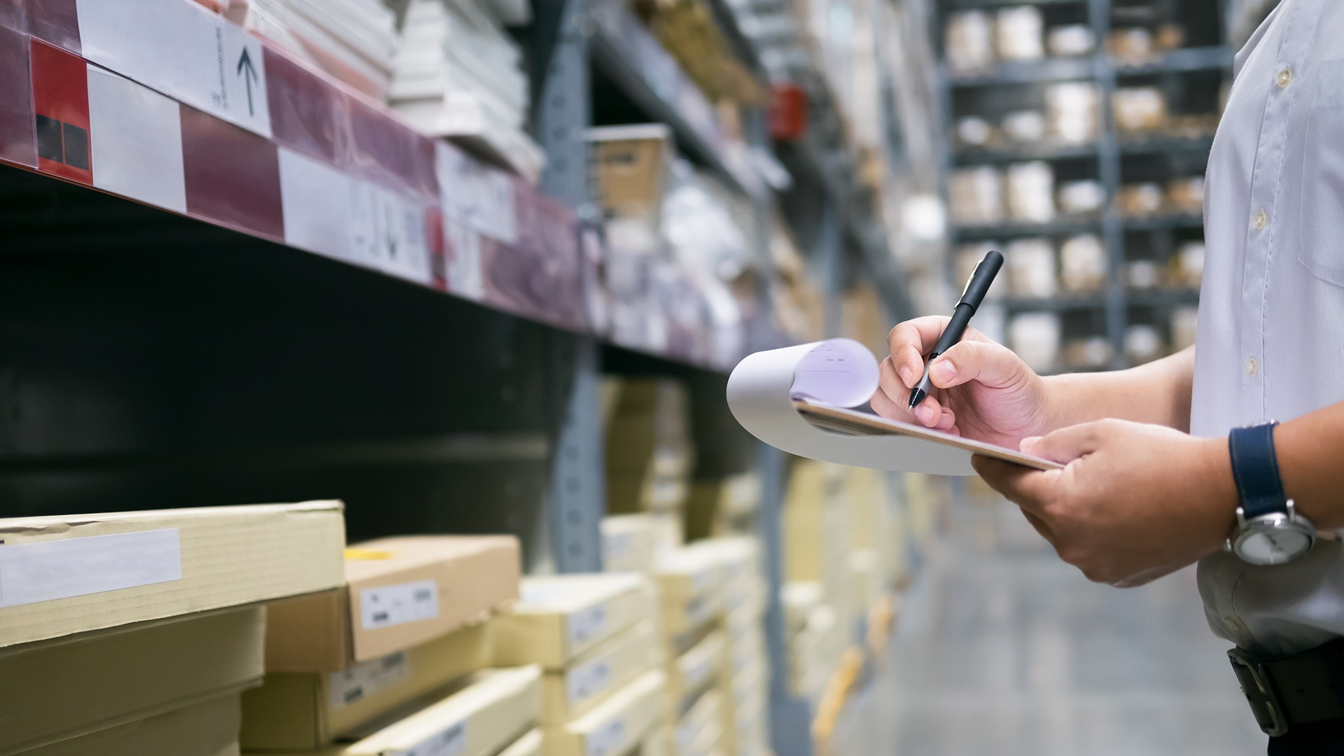 A person inspects shelves filled with boxes in a warehouse, writing on a clipboard. The scene suggests organization and inventory management.