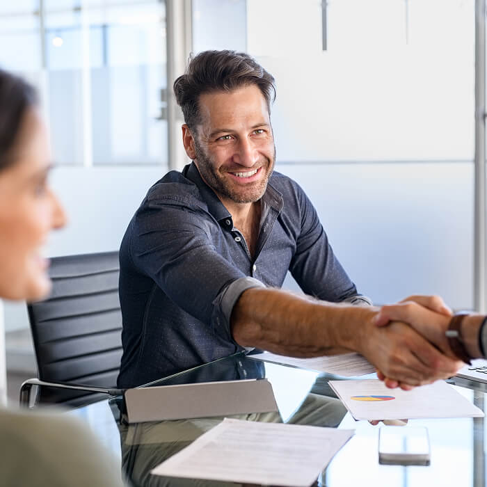 Smiling man in a casual shirt shakes hands across a glass table in a bright office, conveying a friendly and professional atmosphere.
