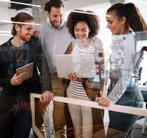 A diverse group of four colleagues smiles while collaborating around a laptop in a modern office, conveying teamwork and positivity.