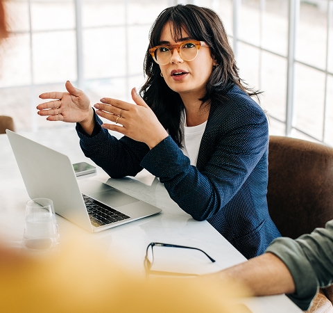 Woman in glasses and blazer gestures while speaking in a meeting, seated at a table with a laptop. The setting is bright and professional.