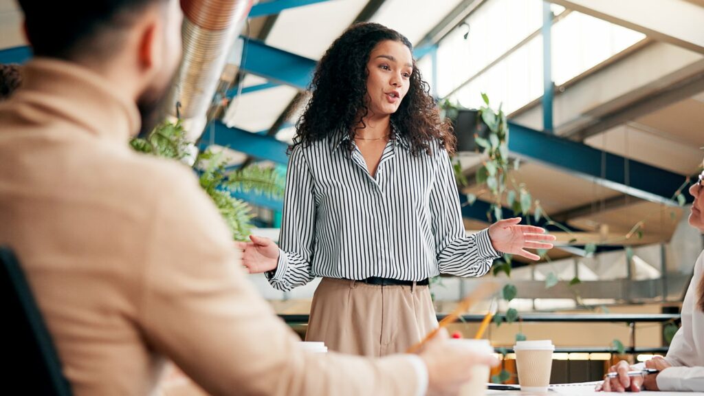 A woman in a striped shirt leads a casual meeting in a modern office, gesturing expressively. Colleagues listen attentively, conveying a collaborative mood.