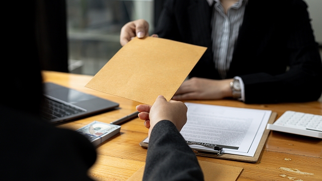 A person hands a brown envelope across a wooden desk to another individual in business attire. A laptop and documents lie nearby, suggesting a professional setting.