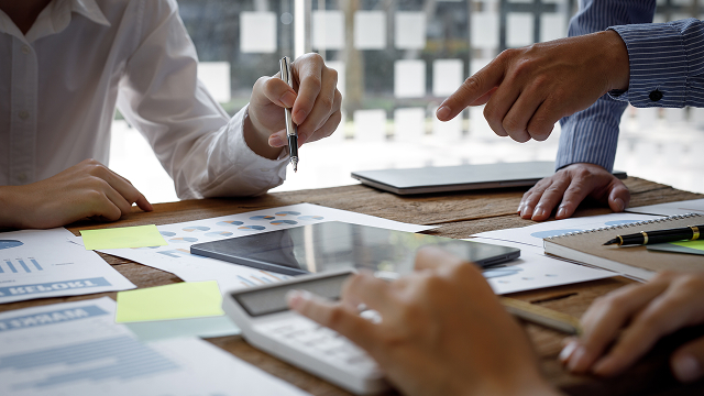 A group of people collaborate around a table filled with documents, a tablet, and a calculator. Hands gesture and point, suggesting an engaging discussion.
