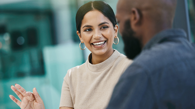 A woman in a tan top with hoop earrings smiles warmly while engaging in conversation with a man in a dark shirt. The setting is bright and modern.