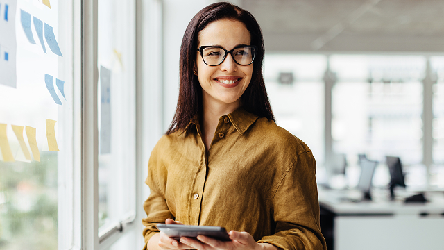 Smiling woman in glasses and a brown blouse holds a tablet in a bright office. Sticky notes adorn the window, creating a productive and cheerful atmosphere.