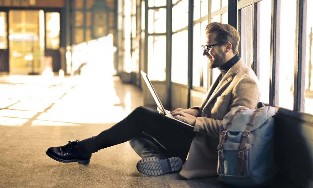 A person seated on the floor, working on a laptop in a sunlit space with large windows, wearing a stylish coat and black shoes.