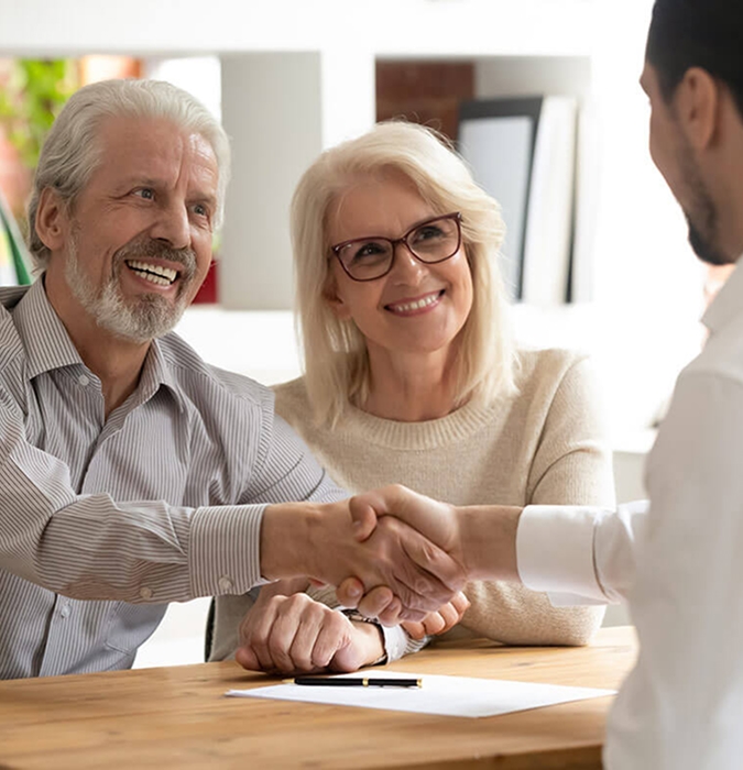 Elderly couple smiles warmly, shaking hands with a person across a table. A document and pen rest on the table, suggesting a positive agreement.