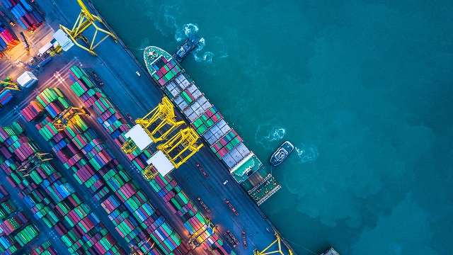 Aerial view of a vibrant port with colorful shipping containers lined up. Cranes stand by a docked ship near turquoise water, evoking a busy industrial vibe.
