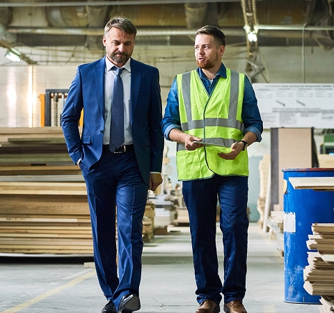 A man in a blue suit and a man in a neon vest walk through a woodwork factory. They appear engaged in conversation, surrounded by wooden planks.