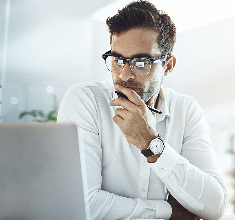 A thoughtful person in glasses, dressed in a white shirt, looks at a laptop while holding a pen to their chin in a bright, modern office space.